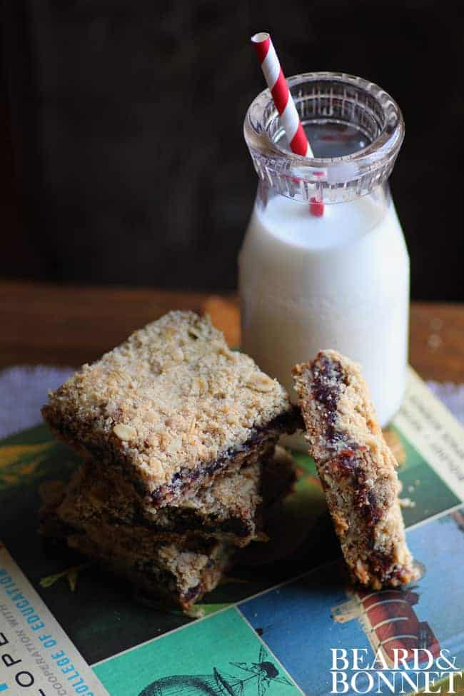 3 date bars stacked on top of one another with a 4th propped next to it. They are all sitting on top of a text book that is green and blue and there is a small bottle of milk with a red and white stripped straw in it next to the book. 