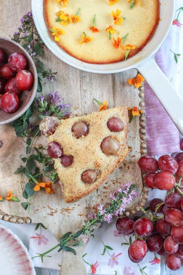 A big wedge of Olive Oil Cake with Polenta and Roasted Red Grapes next to a dish of baked ricotta and a bowl of fresh grapes