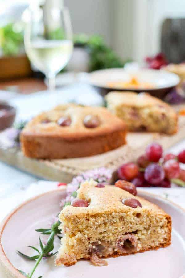 A slice of Olive Oil Cake with Polenta and Roasted Red Grapes recipe on a plate next to a glass of wine