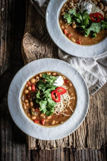 2 bowls of Chickpea and White Bean Chili on table garnished with cheese, cilantro leaves, spur cream, and sliced peppers