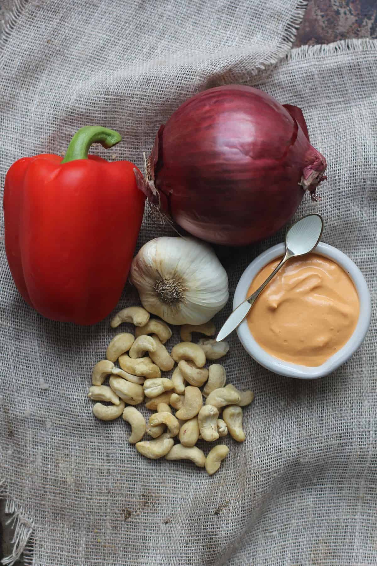 A red bell pepper, cashews, garlic, and red onion are arranged on a piece of burlap. There is a small bowl of cashew cream sauce next to them. 