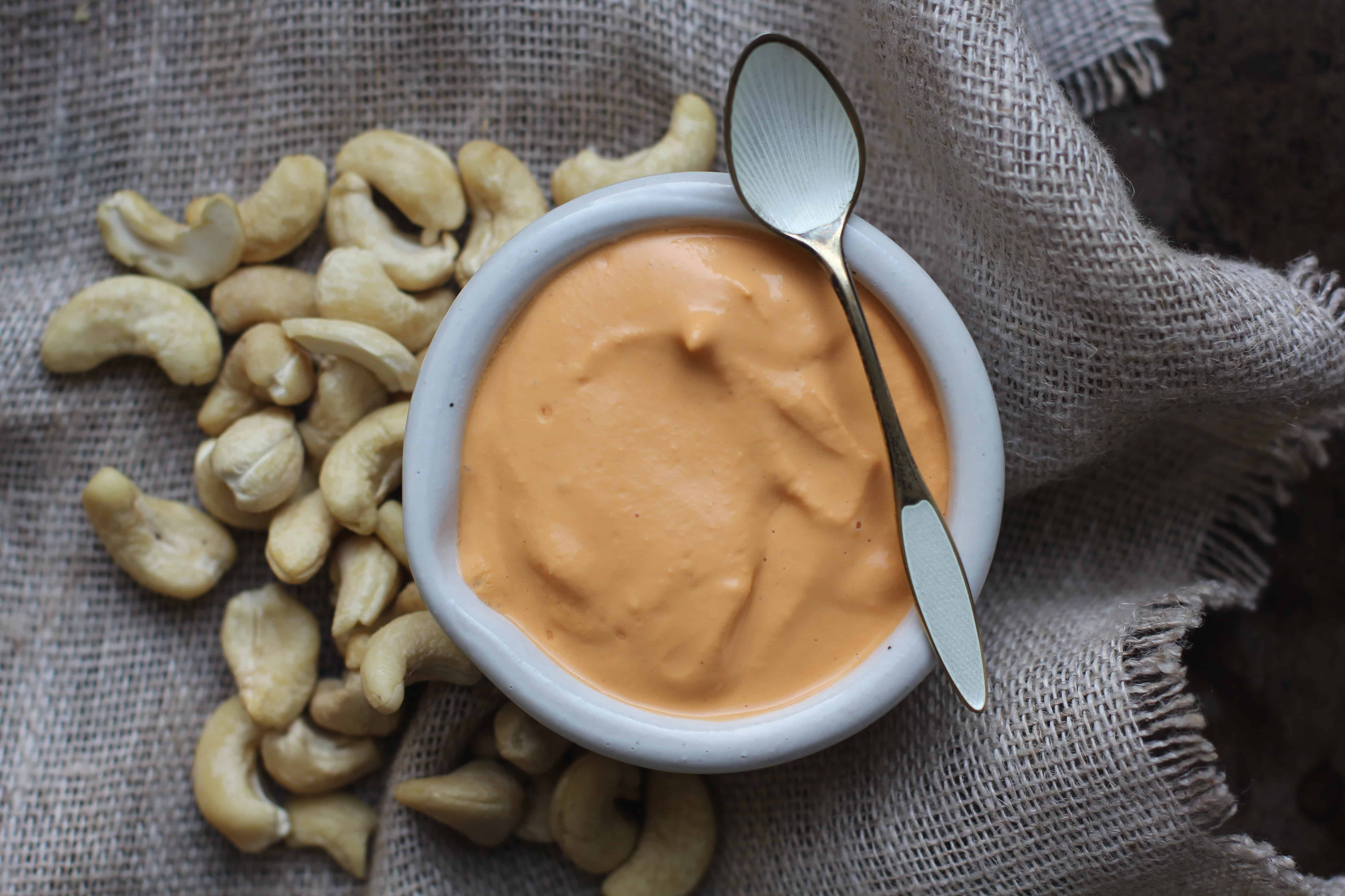 A small white bowl filled with roasted red pepper cashew cream sits on a piece of burlap and has cashews scattered around it.