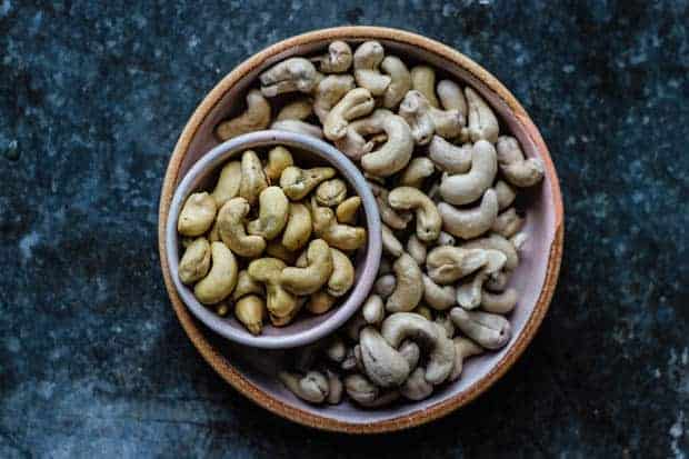 A plate of soaked raw cashews is on a metal table top. there is a smaller bowl of unsoaked raw cashews nestled into th eplate to demonstrate the difference in size