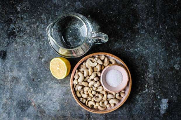 A small glass pitcher of water sits next to alemon half and a plate of cashews on a metal table