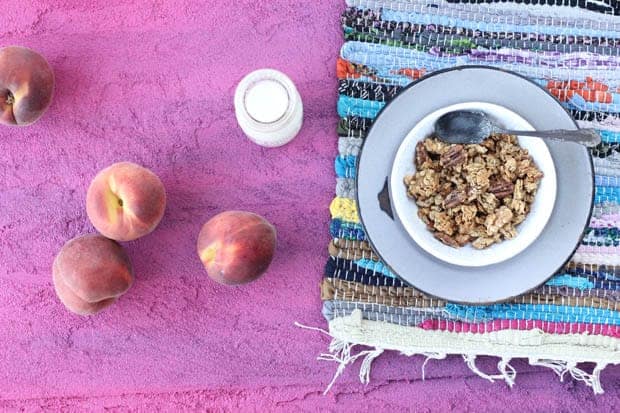 A white bowl of granola on a gray enamelware plate on top of a multi-colored woven placemat. The placemat is on top of a magenta colored table with a clear glass jug of milk and 4 fresh peaches on top.