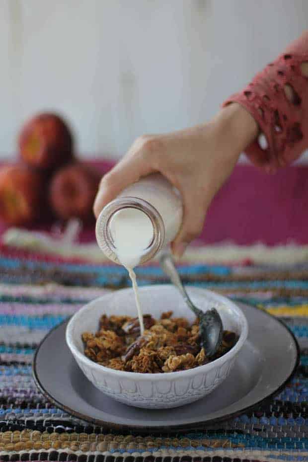 A white bowl of granola is sitting on top if a gray enamelware plate on a multi-colored woven placemat. There is a woman's hand pouring milk from a clear glass milk jug into the granola. The woman is wearing a coral colored long sleeve shirt with a decorative edge around the cuff. The table underneath the placemat is magenta and there are 3 peaches stacked up in the background. 