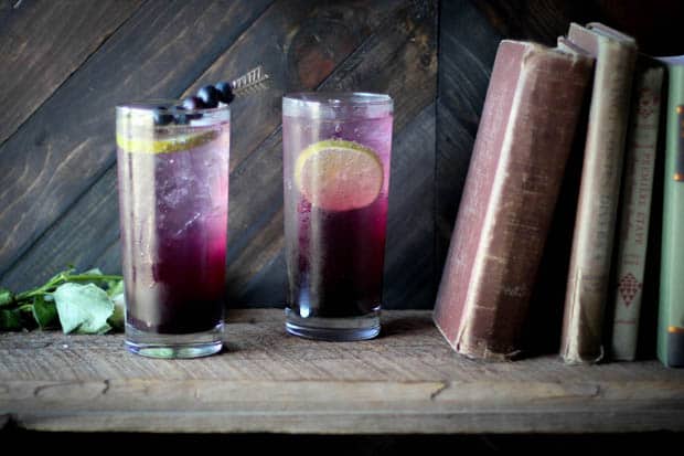 2 tall clear glass filled with ice and a purple ombre drink on a wooden book shelf next to 4 vintage books that are leaning to the right side of the image. 