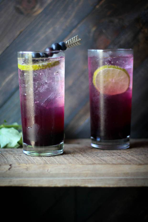 2 tall glasses sitting on a wooden shelf. They are filled with ice and a drink that has a purple ombre effect. There are lime wheels in both drinks and one of them has a cocktail pick with blueberries threaded onto it laying across teh top. There is a wall in the background made of dark brown wood slats. 