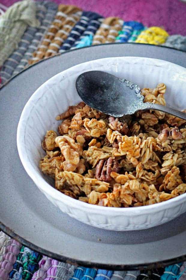 Peach cobbler granola in a white bowl is sitting on top of a gray enamelware plate. The plate is on a multicolored woven placemat that is on top of a magenta colored table. 