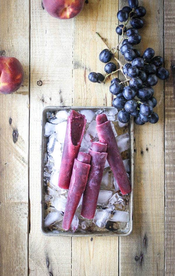 Grape and peach ice pops on a tray of ice. There are peaches and grapes on the table top next to the grapes