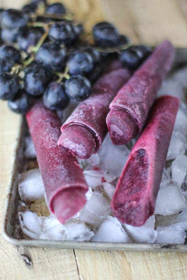 4 purple ice pops sitting on a small tray with ice. A cluster of grapes are in the background. 