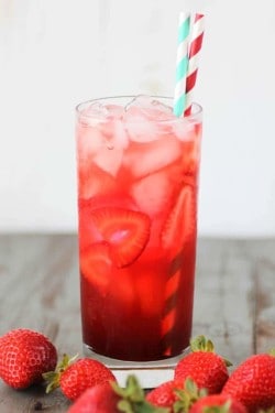 A glass of pink strawberry soda is on a table - it has a white and blue and a red and white straw. There are also whole berries on the table.