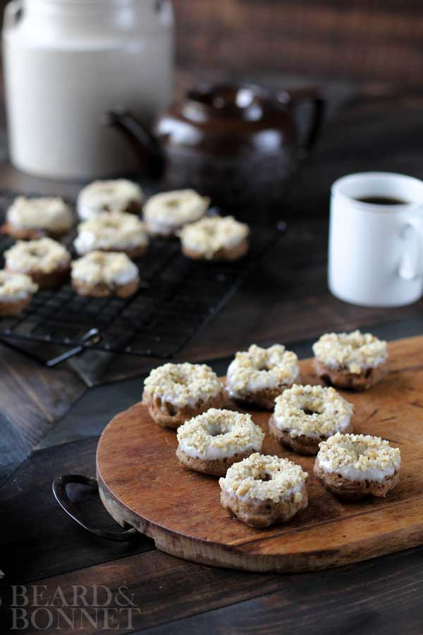 7 mini banana bread donuts on a wood cutting board with more mini donuts on a wire rack behind it. A white coffee mug filled with coffee is next to the cutting board as well as a brown tea pot