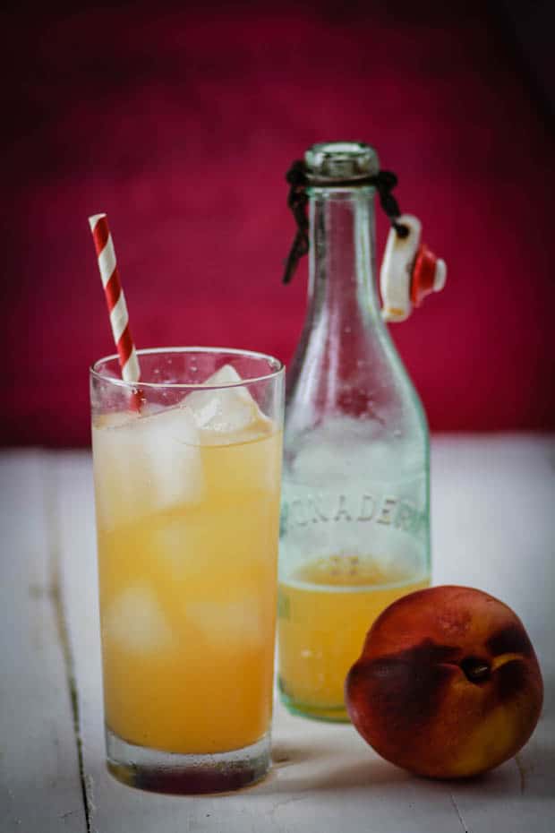 A glass of peach and ginger soda with a red and white straw sits next to a bottle of soda syrup and a fresh peach. The background of the image is magenta. 