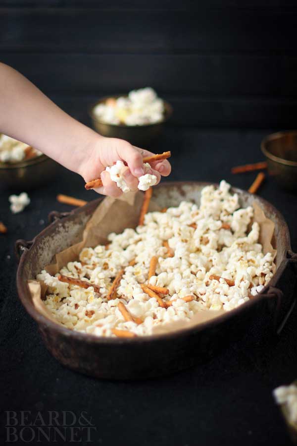 A little kids hand is in the image grabbing a handful of the spooky popcorn mix. It has popcorn, candy corn, pretzel sticks, and candy eyeballs