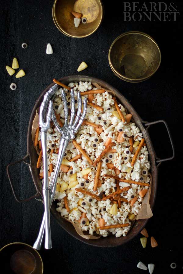 Sppoky popcorn mix with popcorn, candy corn, pretzel sticks, and candy eyeballs in an oval serving dish with skelteon hands serving spoons. 