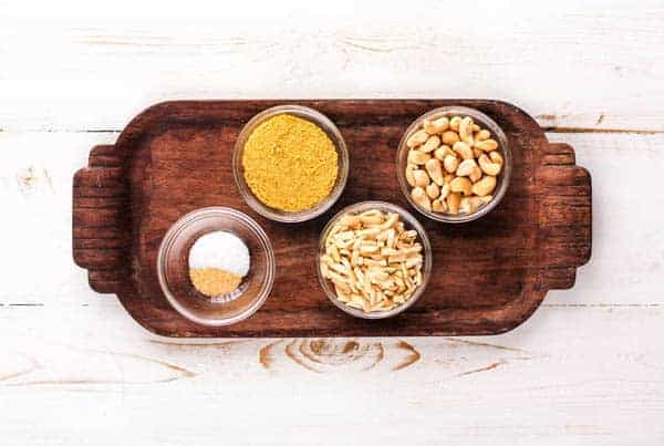 A small wooden tart with bowls of ingredients. Cashews, nutritional yeast, almonds, salt, and garlic powder