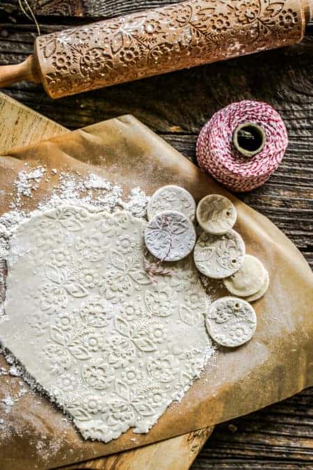 A baking scene with salt dough rolled out on parchment the imprinted with a design. Next to the dough are a few finished salt dough ornaments a spindle of red and white string and a rolling pin that has a relief pattern carved in the dough. 