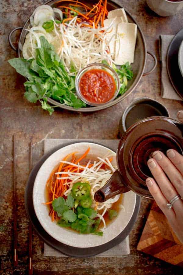 a woman pouring pho broth over a bowl of noodles and pho toppings