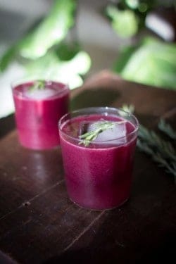 2 Stunning purple Rosemary Concord Grape Cocktails on a table. The cocktails are styled with 1 large square ice cube and a sprig of fresh rosemary.