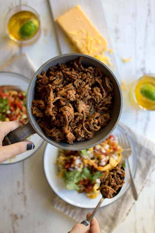 A skillet of Gluten Free Taco Spiced Ground Beef, a woman is spewing beef into assembled taco bowls