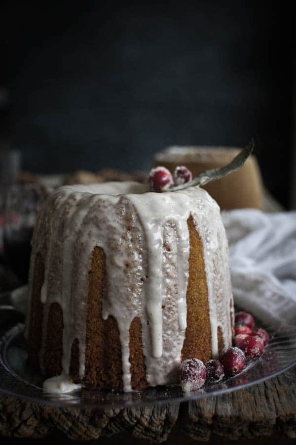 A bundt cake on a clear plate drizzled with eggnog icing and topped with sugared cranberries and bay leaves