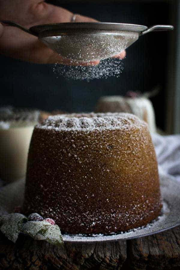 A tall bundt cake is on a clear plate. There is powdered sugar being sifted over the top of the cake. 