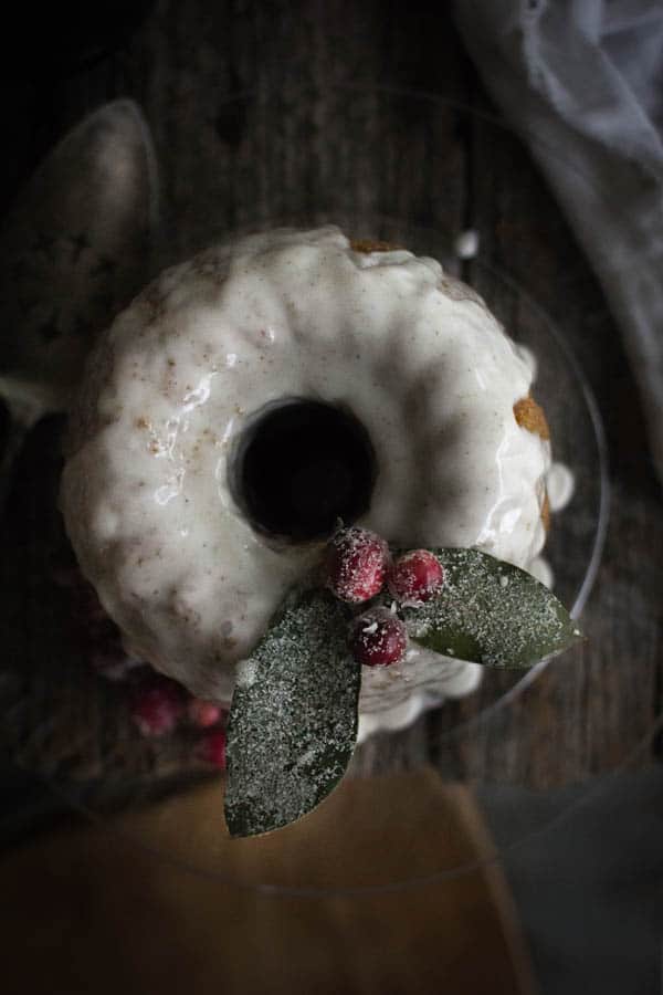 looking down on top of a bundt cake glazed in eggnog icing. There are sugared bay leaves and cranberries on top that look like holly