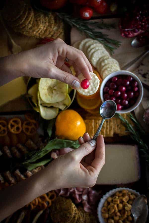 A woman hands holding a cracker with a bright red pickled cranberry on top over a very large fall inspired cheese board