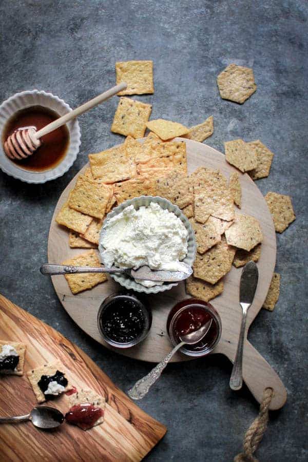 a wooden food board with homemade ricotta, strawberry jam, and blueberry jam and crackers. 