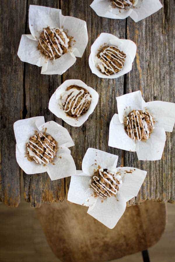 7 Iced Cinnamon Oat Cakes are sitting in their white parchment baking liners on an old wooden table top. There is a wooden seat in the bottom right hand corner of the image that is pushed up close to the table. The oat cakes are brown and the drizzle of icing is white.