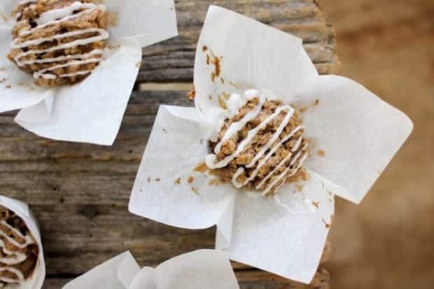 There are 3 iced cinnamon oat cake with white icing drizzle is sitting on top of its white muffin liner that has been peeled back. They are sitting on a wooden table top.