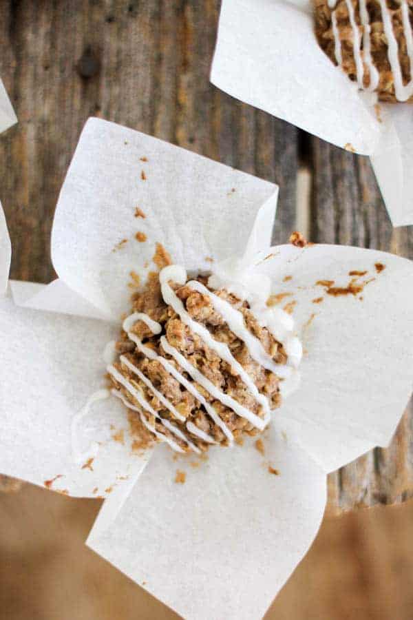 A cinnamon oat cake with white icing drizzle is sitting on top of its white muffin liner that has been peeled back. They are sitting on a wooden table top. There is another oat cake in teh upper right hand corner of the frame. 