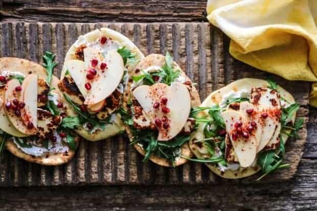 flatbread topped with labneh, arugula, pan seared halloumi, apple, pomegranate, aleppo pepper on a wooden board on a weathered wooden table top with a yellow napkin