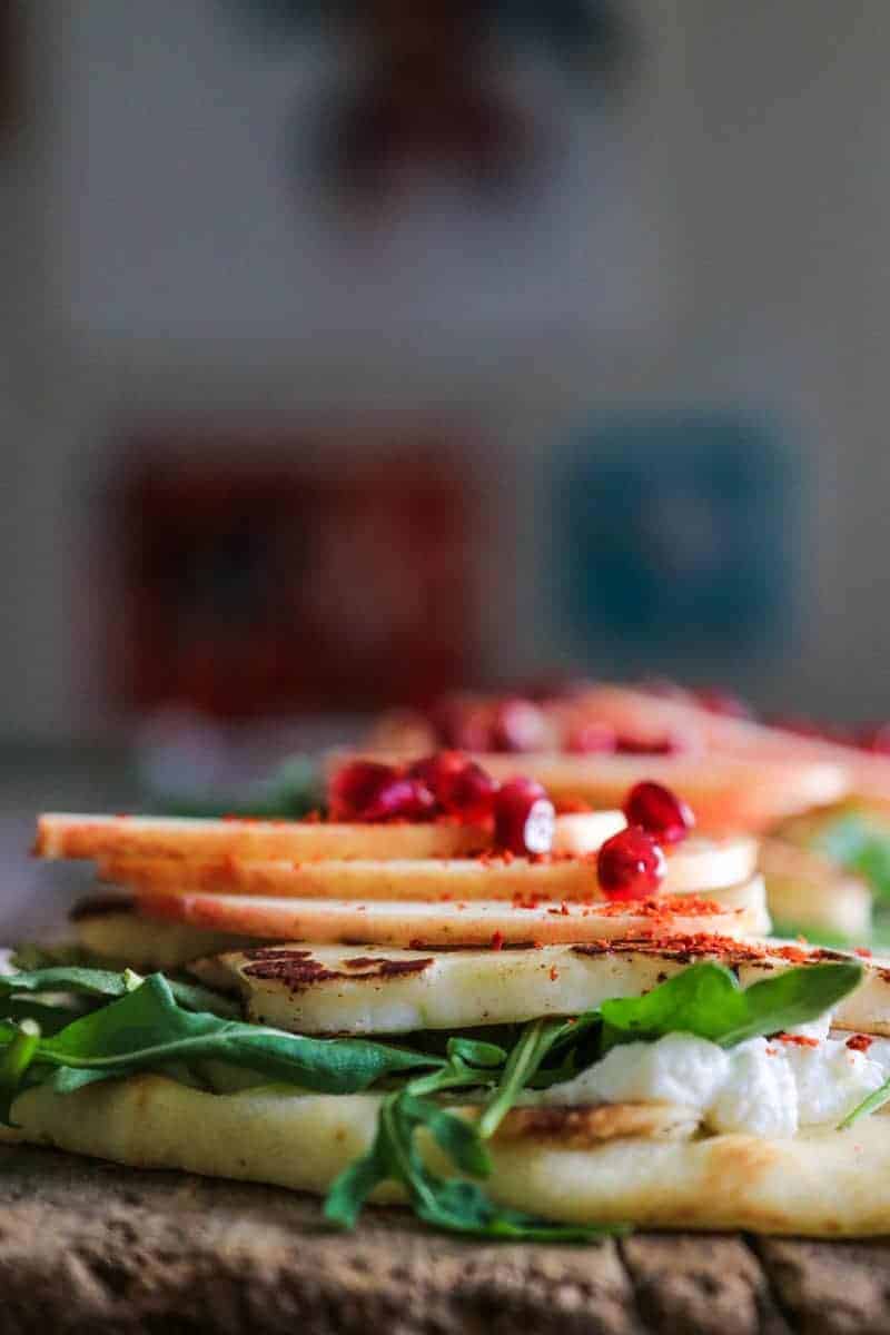 Flatbread layered with labneh, arugula, pan seared halloumi cheese, apple slices, aleppo pepper, and pomegranate