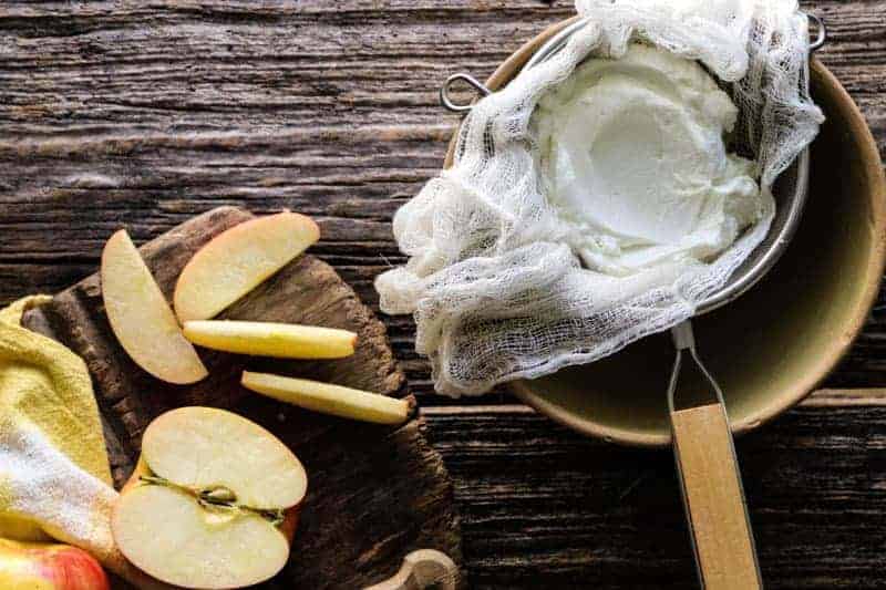 Greek yogurt in a strainer lined with cheesecloth next to a platter with sliced apples.