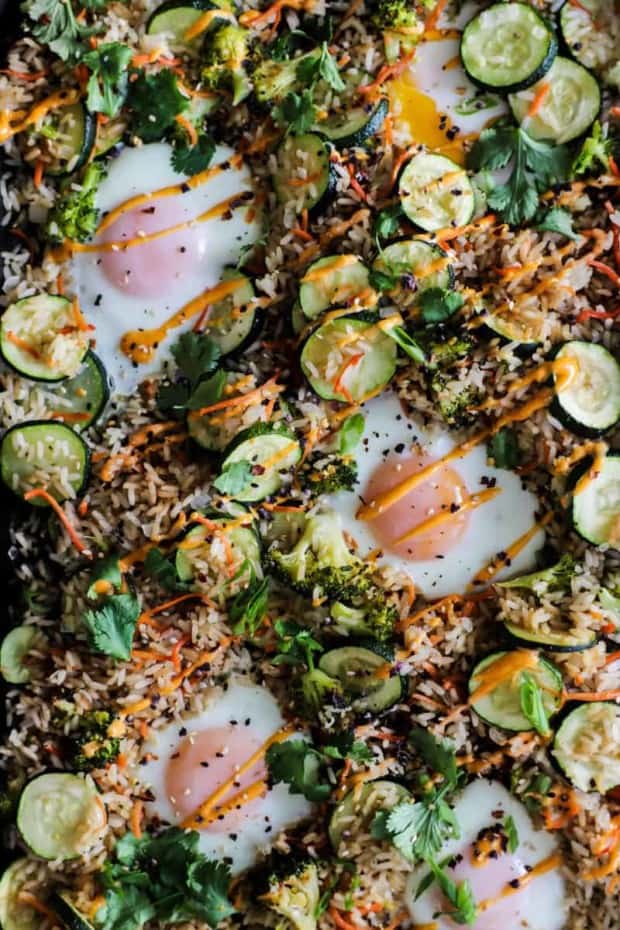 sheet pan full of oven fried rice, zucchini sliced, carrot, broccoli, and herbs with eggs and a sriracha mayo drizzle.