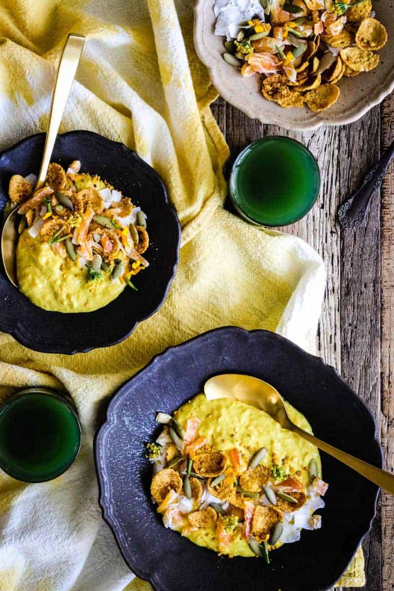 2 bowls of yellow golden milk yogurt, 2 glasses of green juice, and a bowl with granola, coconut flakes, pepitas, and chopped dried mandarins.