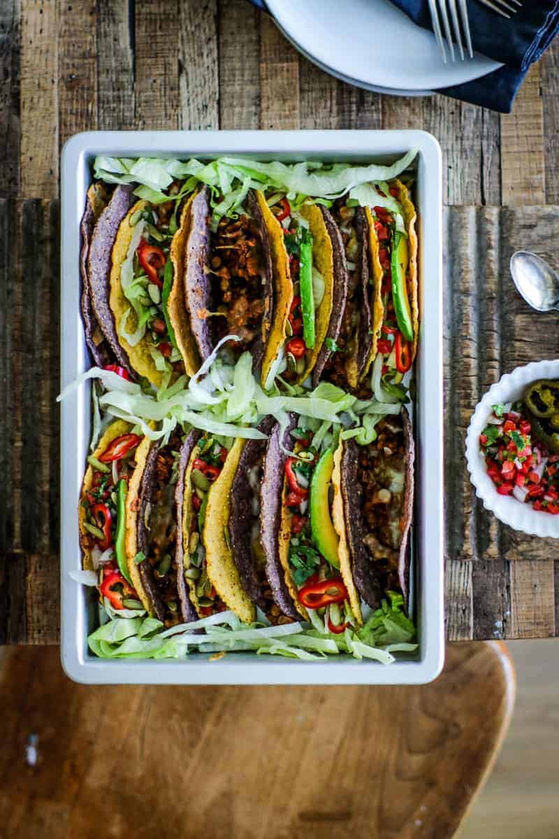 Baking pan filled with tacos made of lentils and meatless beef crumbles topped with lettuce, pico de gallo, peppers, avocado slices, and pepitas