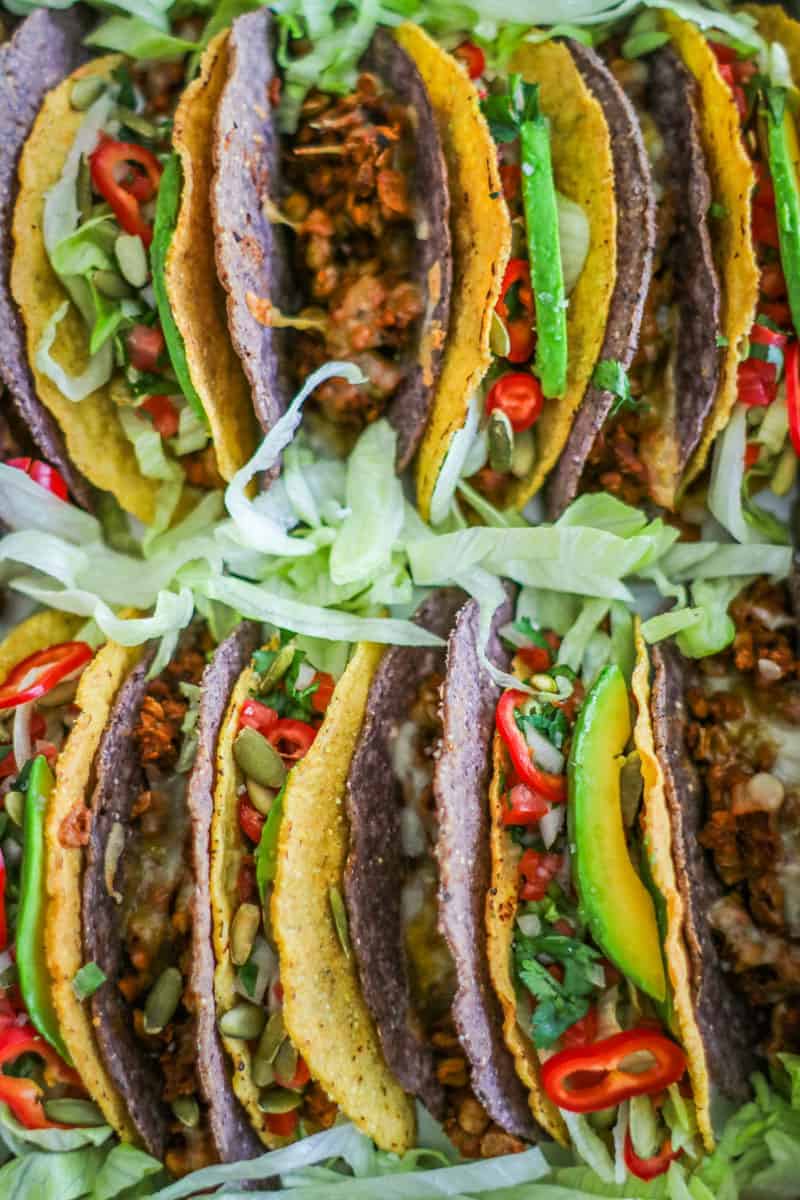 close up of Baking pan filled with tacos made of lentils and meatless beef crumbles topped with lettuce, pico de gallo, peppers, avocado slices, and pepitas