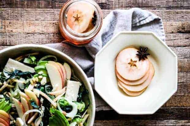 Salad in an off white stone ware bowl made of greens, fresh sliced apples, shaved fennel, chopped pickled apples, and white cheddar cheese chunks on a tobacco lathe table top with an octagonal shaped off white bowl filled with slices of pickled apples and a star anise on top of a linen napkin. There is also a mason jar filled with slices of pickled apples.