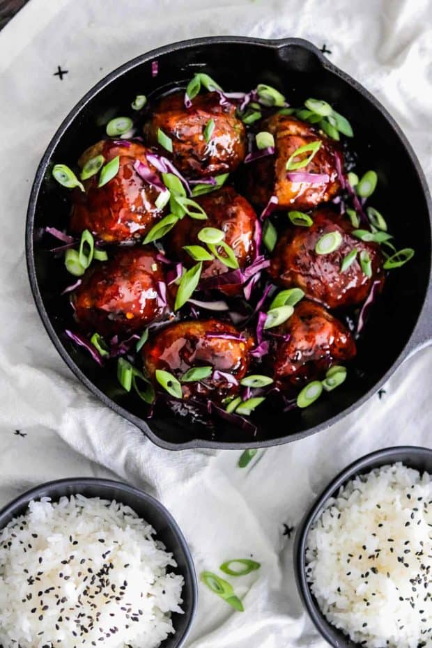 Korean BBQ Meatballs in a skillet with green onions and red cabbage. @ bowls of rice on the side with black sesame seeds.