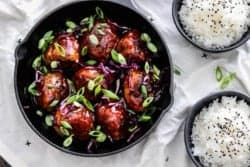 Korean BBQ Meatballs in a skillet with green onions and red cabbage. @ bowls of rice on the side with black sesame seeds.