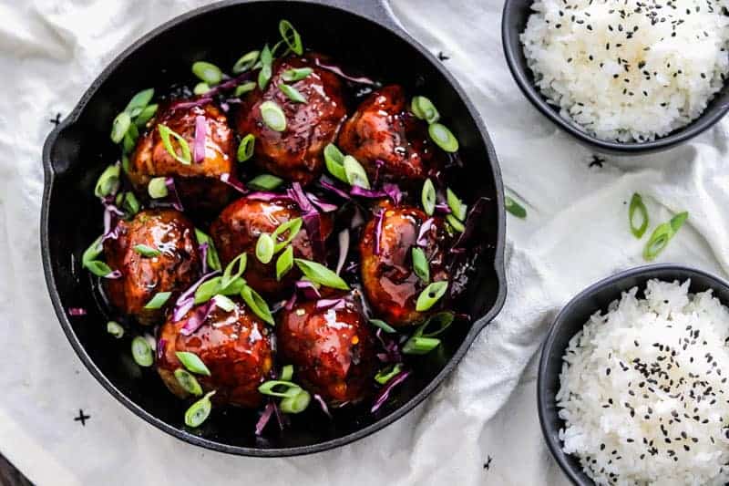 Korean BBQ Meatball Recipe in a skillet with green onions and red cabbage. @ bowls of rice on the side with black sesame seeds.