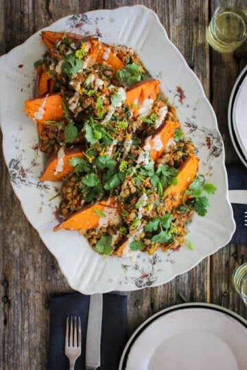 A large rectangular platter is lined with roasted sweet potatoes halves which are topped with spiced lentils, lemon tahini dressing, and fresh cilantro. Teh platter sits on a weathered wooden table along with 2 place settings.