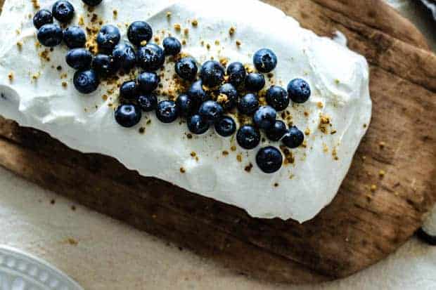 looking down onto a rectangular cake that is frosted with white whipped cream icing and sprinkled with fresh blueberries and yellow bee pollen. Cake is on a wooden board with a metal handle on a light colored table.