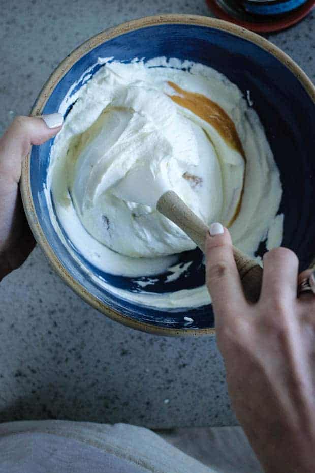 A woman holding a vintage blue mixing bowl with an off-white rim over a gray quartz countertop. She is using a white, rubber spatula to fold lemon curd into whipped cream