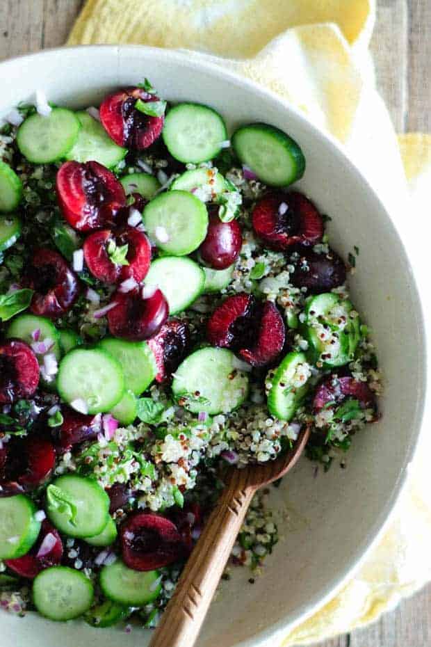 A cream colored bowl filled with halved cherries, quinoa, cucumbers, and basil. On top of a wooden table with a yellow napkin.