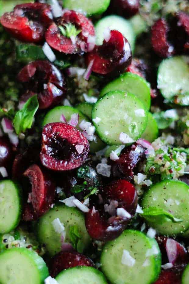 A close up image of Cherry Basil Tabbouleh with halves of deep red cherries, thin slices of Persian cucumber, finely minced red onion, tricolored quinoa, and basil leaves.