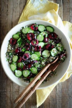 A cream bowl placed on top of a yellow and white cloth napkin on a tobacco lathe table top. The bowl is filled with tabbouleh made with quinoa, cucumber rounds, halved cherries, and basil. There are 2 wooden serving utensils in the bowl.